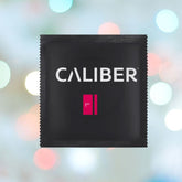 A black square packet with "Caliber L - Large Condoms (57mm Wide)" in magenta, placed against a background of blurred multicolored lights.